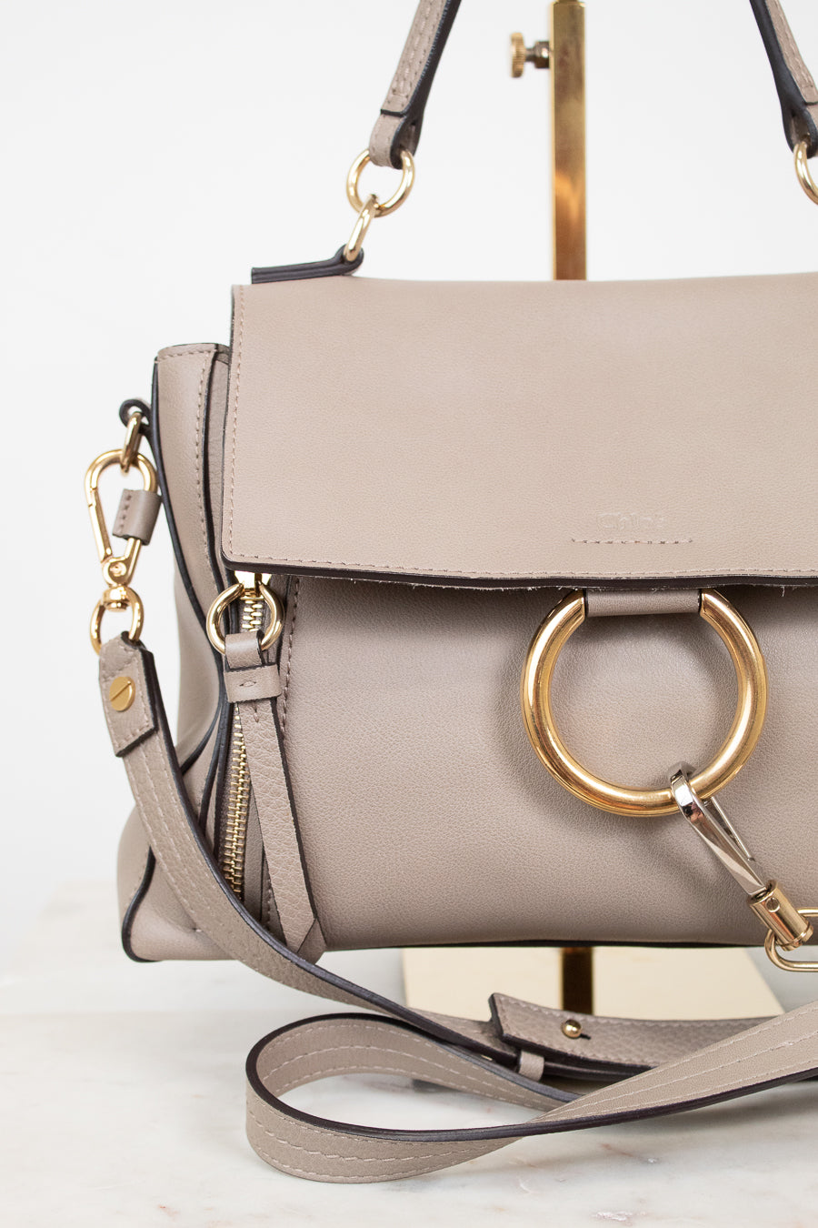 A taupe leather handbag with a large gold-tone O-ring on the front flap, a matching shoulder strap, and gold-tone hardware, displayed on a stand against a white background.