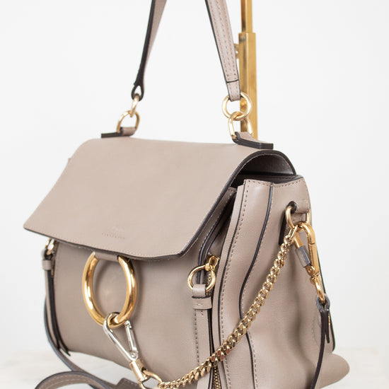 A taupe leather handbag with gold-tone hardware, featuring a flap top, a gold-tone O-ring, a gold chain strap, and a structured rectangular handle, shown on a white background.