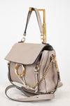 A taupe leather handbag with gold-tone hardware, featuring a flap top, a gold-tone O-ring, a gold chain strap, and a structured rectangular handle, shown on a white background.