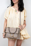 A woman wearing a pale yellow and white gingham dress carries a taupe leather shoulder bag with gold-tone hardware and a large circular ring detail on the front.