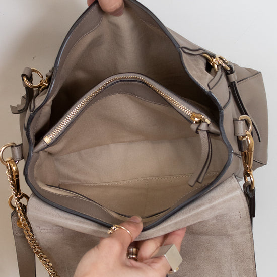 A taupe suede shoulder bag with gold hardware, shown open with hands holding the top and interior compartments visible, including a zippered pocket.