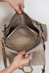 A taupe suede shoulder bag with gold hardware, shown open with hands holding the top and interior compartments visible, including a zippered pocket.