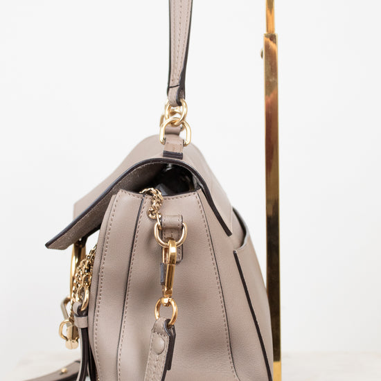 A taupe leather handbag with gold-tone hardware, shown from a side angle resting on a gold metal display stand against a white background.