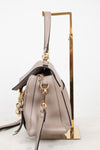 A taupe leather handbag with gold-tone hardware, shown from a side angle resting on a gold metal display stand against a white background.
