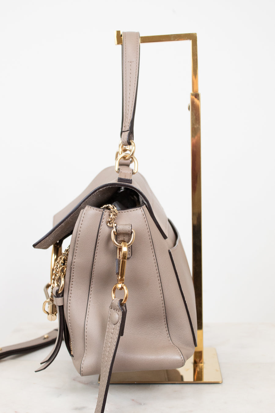 A taupe leather handbag with gold-tone hardware, shown from a side angle resting on a gold metal display stand against a white background.