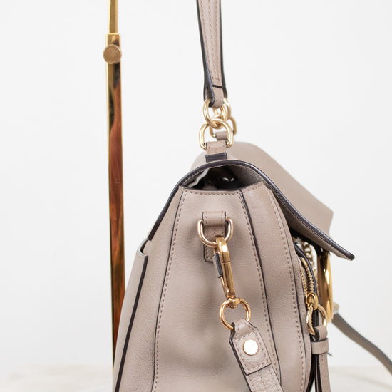 A taupe leather handbag with gold-tone hardware, shown from the side on a gold display stand against a white background.