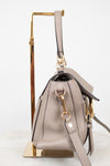 A taupe leather handbag with gold-tone hardware, shown from the side on a gold display stand against a white background.