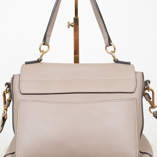 A structured taupe leather handbag with a top handle and gold-tone hardware, displayed on a gold stand against a white background.