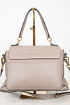 A structured taupe leather handbag with a top handle and gold-tone hardware, displayed on a gold stand against a white background.