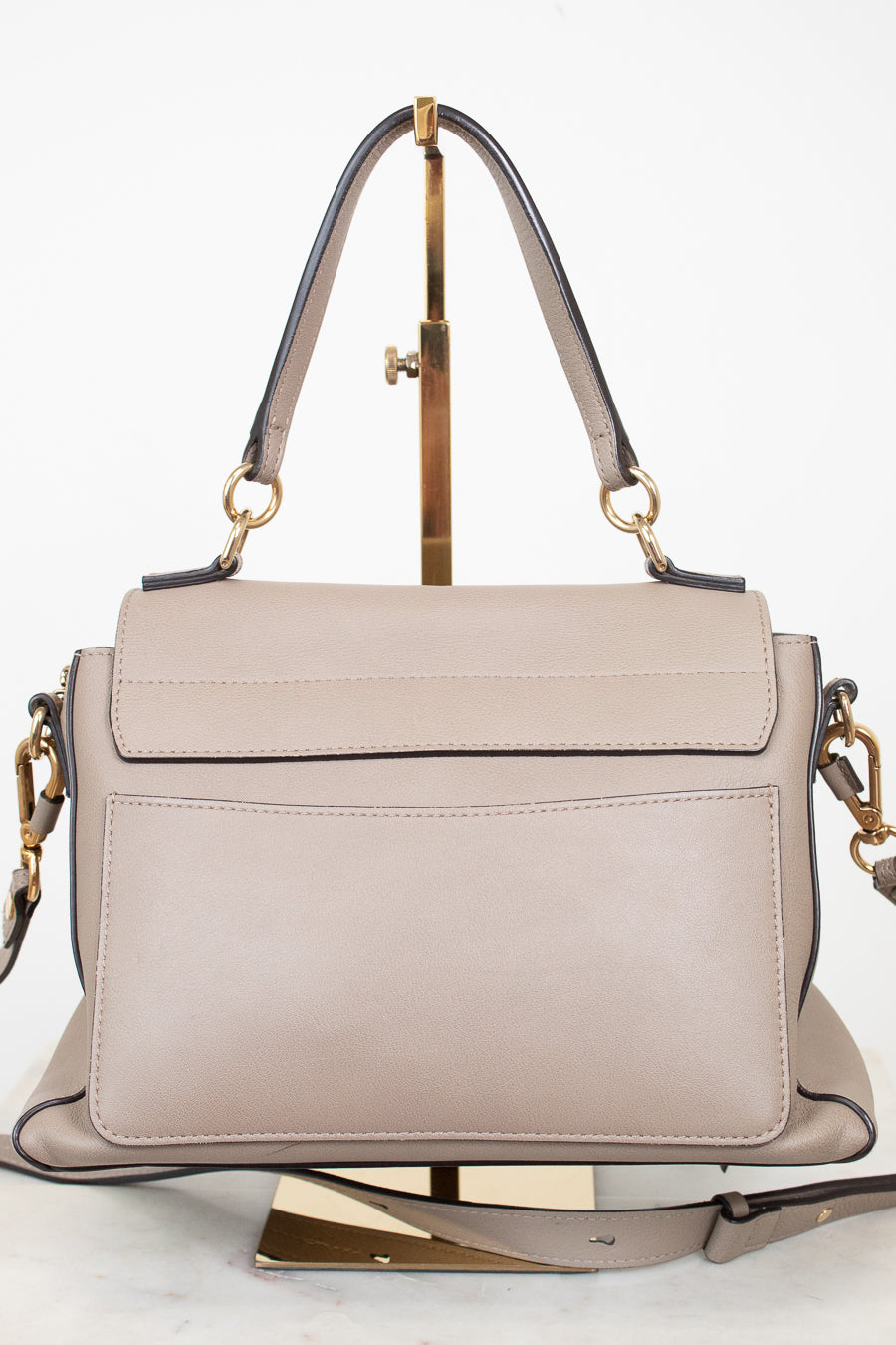 A structured taupe leather handbag with a top handle and gold-tone hardware, displayed on a gold stand against a white background.