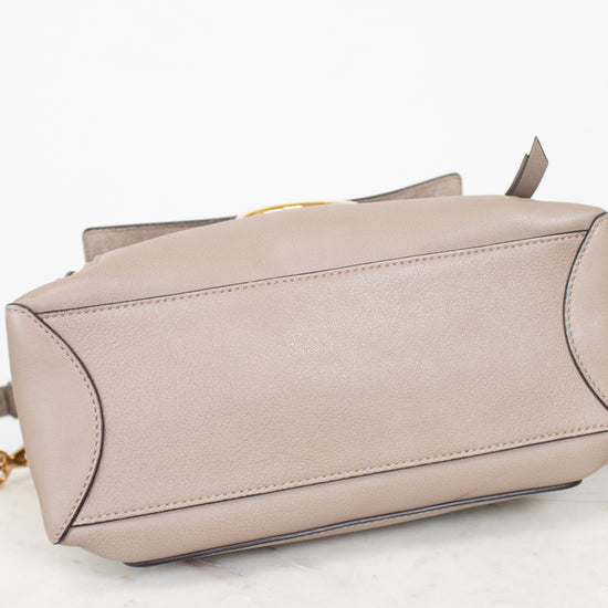 A taupe leather handbag shown from a side angle, resting on a white marble surface against a white background.