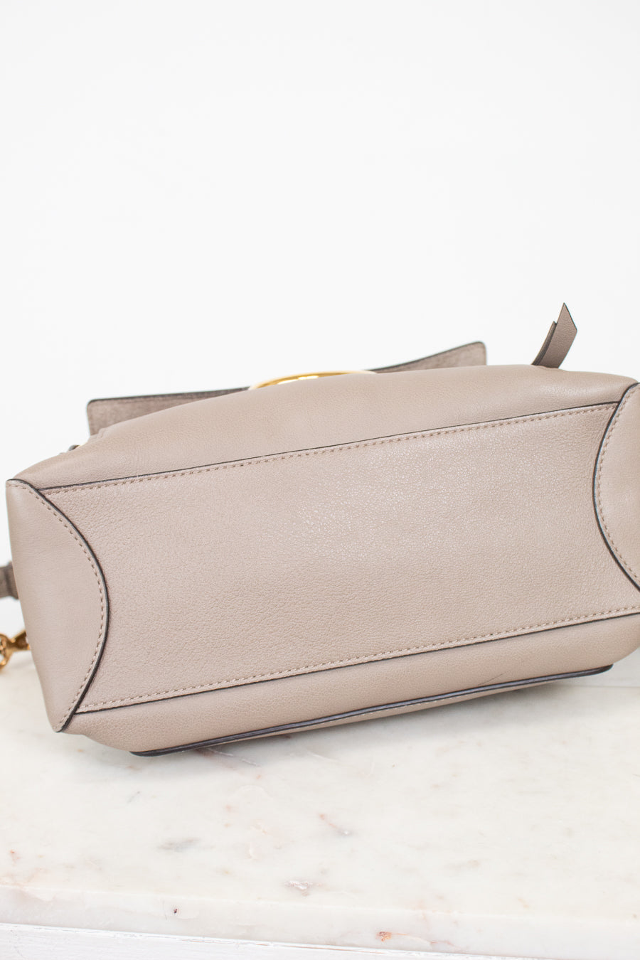 A taupe leather handbag shown from a side angle, resting on a white marble surface against a white background.
