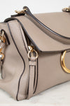 A close-up of a taupe leather handbag with gold-tone hardware, featuring a flap closure, top handle, and a prominent circular gold ring detail on the front.