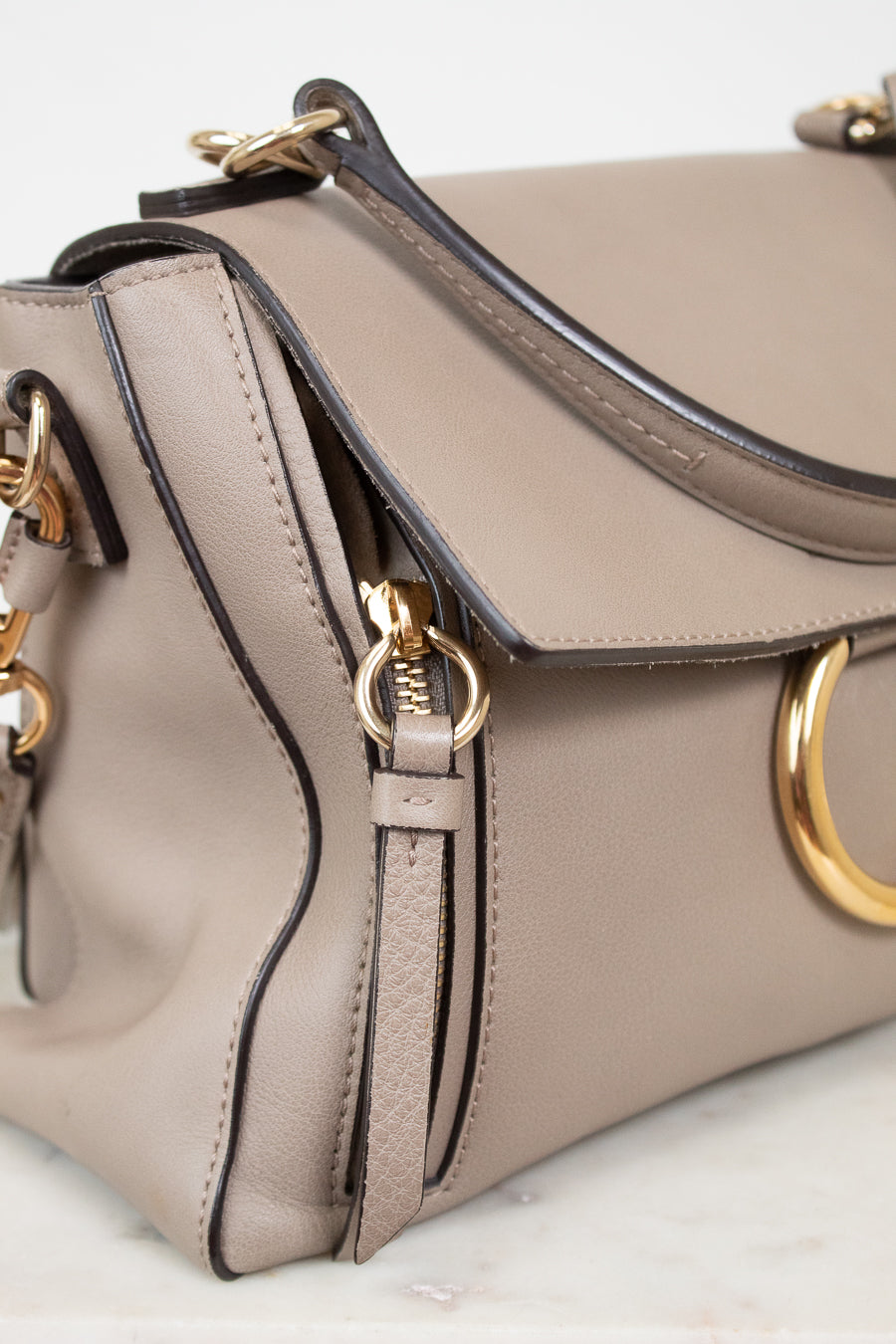 A close-up of a taupe leather handbag with gold-tone hardware, featuring a flap closure, top handle, and a prominent circular gold ring detail on the front.