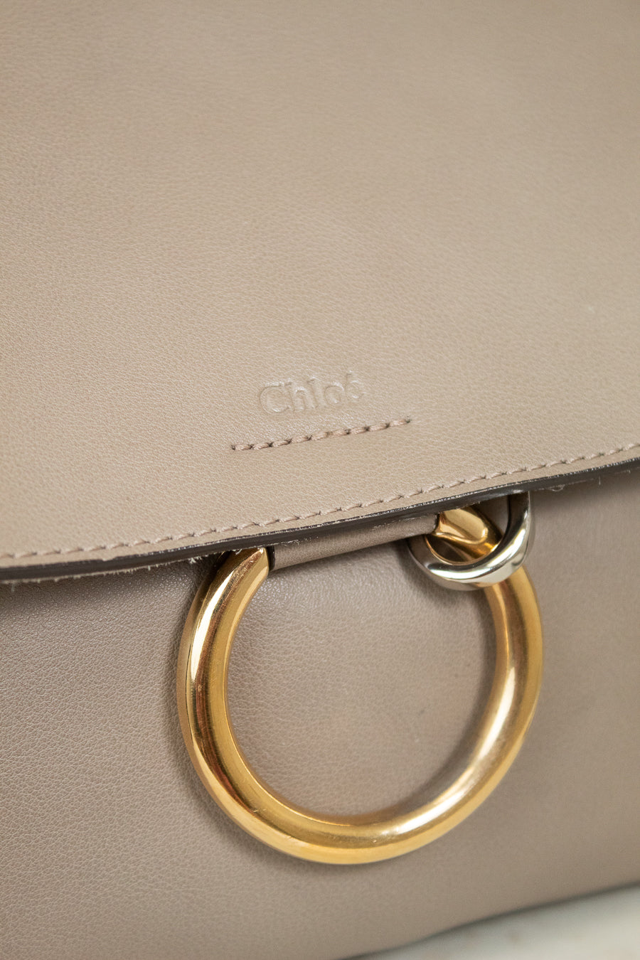 Close-up of a taupe leather handbag with a gold-tone O-ring handle and an embossed Chloé logo on the flap.