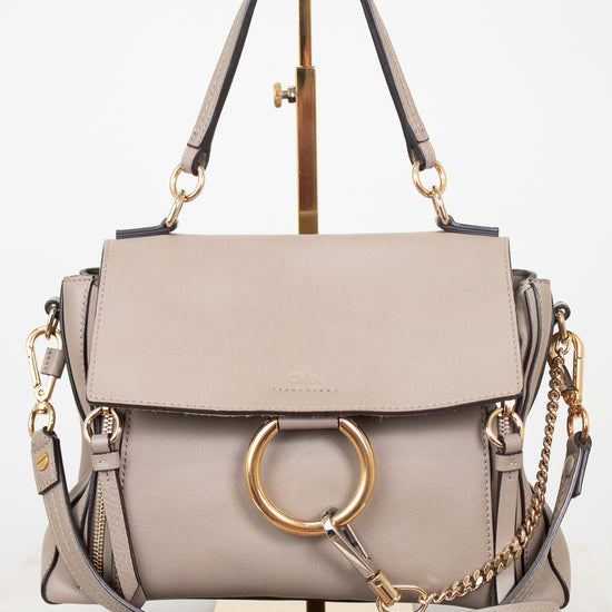 A structured taupe leather handbag with a gold-tone circular ring detail, top handles, and a detachable shoulder strap, displayed on a stand against a white background.