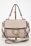 A structured taupe leather handbag with a gold-tone circular ring detail, top handles, and a detachable shoulder strap, displayed on a stand against a white background.