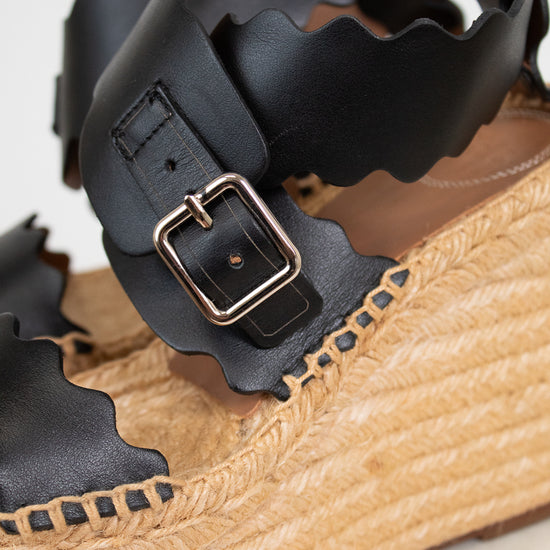 Close-up of a sandal with black leather straps and a tan woven sole.