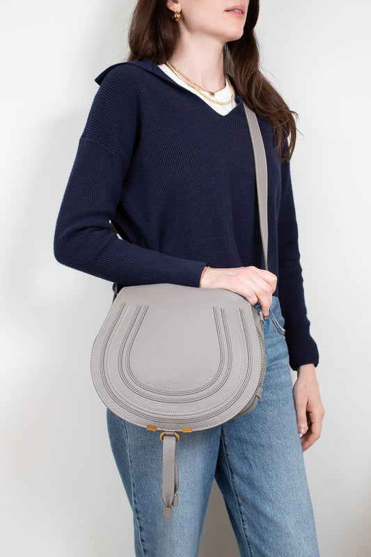 Person wearing navy sweater and jeans holding a grey saddle crossbody bag on a white background