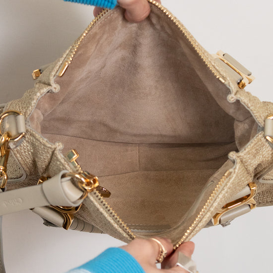 Close-up of a beige handbag with gold accents held by a person wearing a blue sweater.