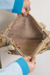 Close-up of a beige handbag with gold accents held by a person wearing a blue sweater.