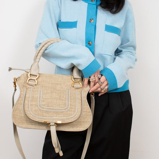 Person wearing a light blue jacket with dark buttons, holding a beige handbag against a white background