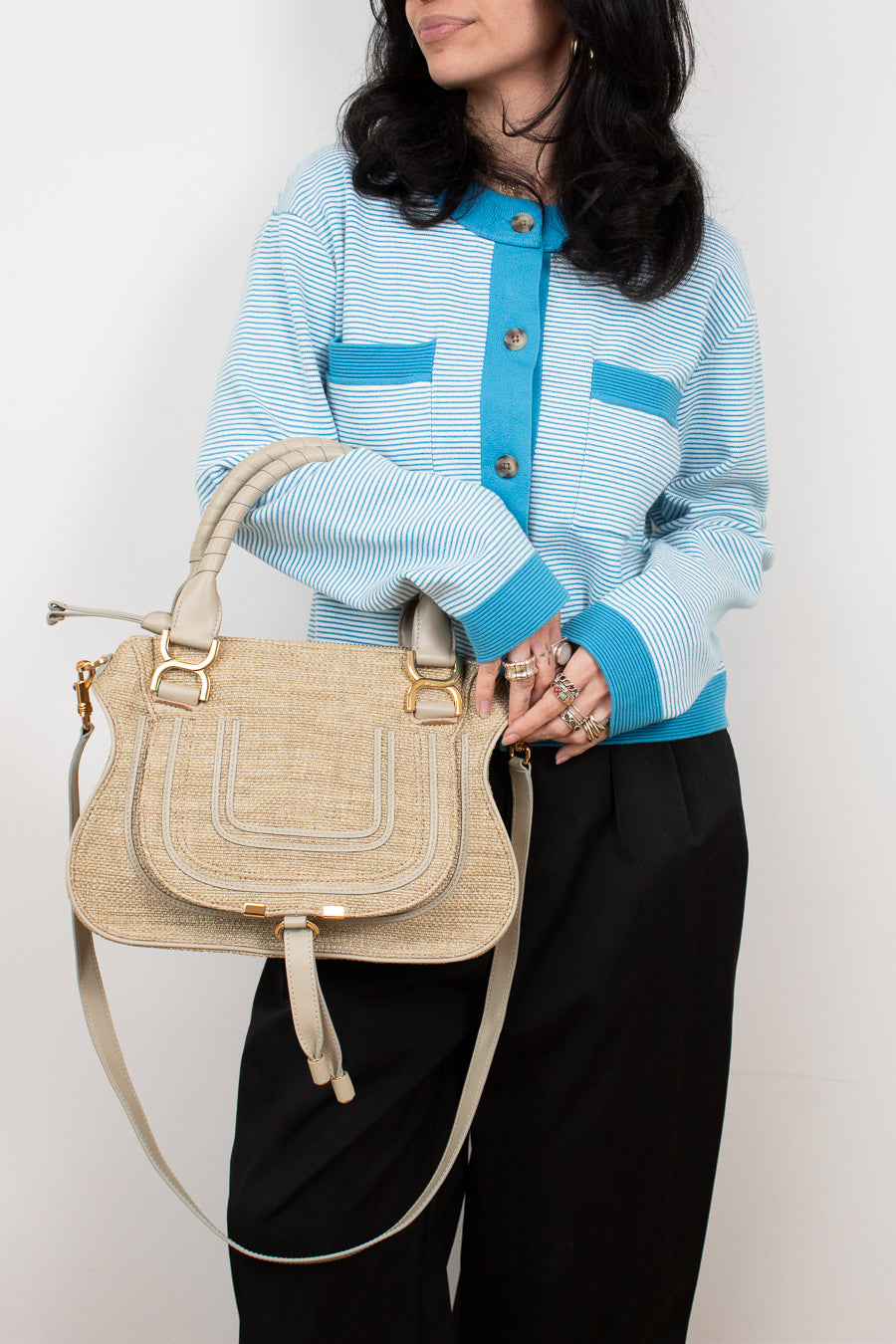 Person wearing a light blue jacket with dark buttons, holding a beige handbag against a white background