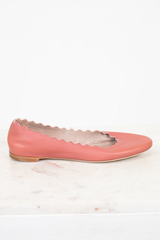 A single dusty pink leather flat shoe with a scalloped cutout edge detail, resting on a white marble surface against a white background.