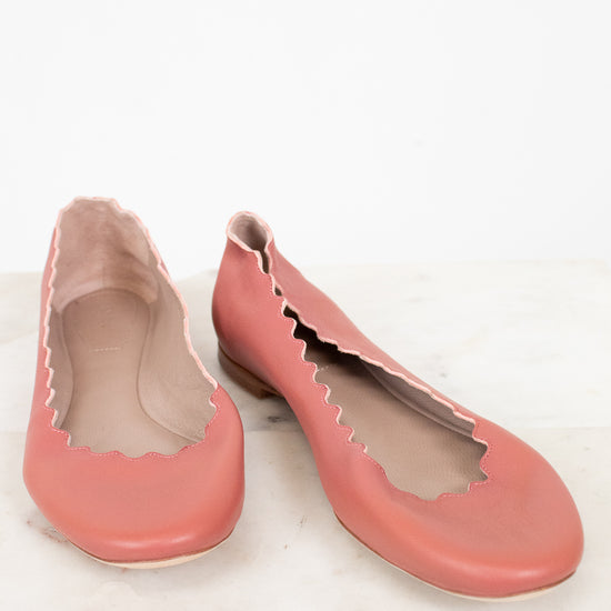 A pair of dusty pink leather ballet flats with scalloped edges, displayed on a light marble surface against a white background.