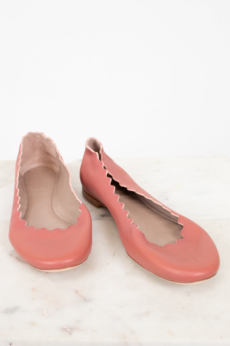 A pair of dusty pink leather ballet flats with scalloped edges, displayed on a light marble surface against a white background.