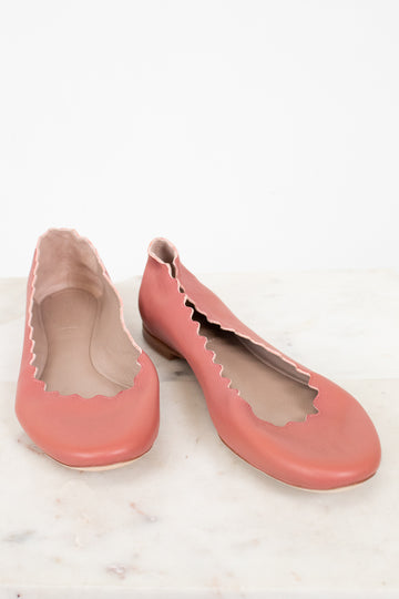 A pair of dusty pink leather ballet flats with scalloped edges, displayed on a light marble surface against a white background.