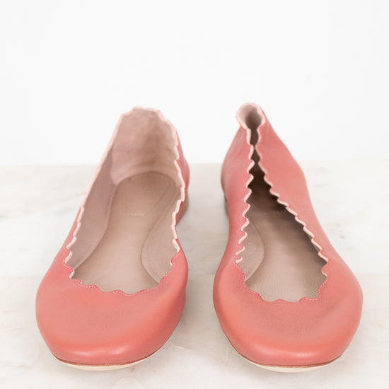 A pair of dusty pink leather ballet flats with scalloped edges, shown from the side on a light marble surface against a white background.