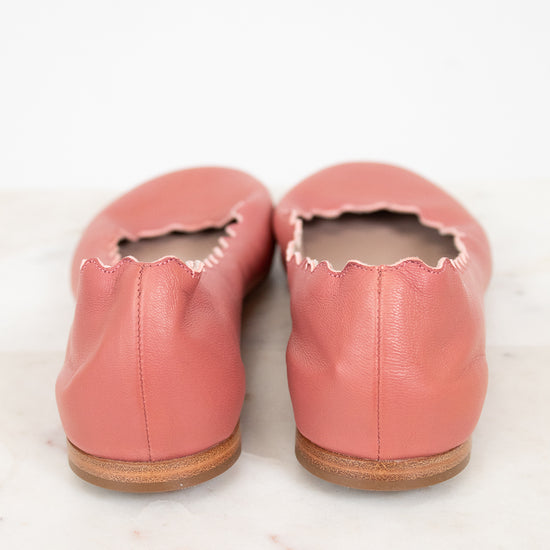 A pair of dusty pink leather ballet flats with scalloped edges, shown from the back on a white marble surface against a white background.