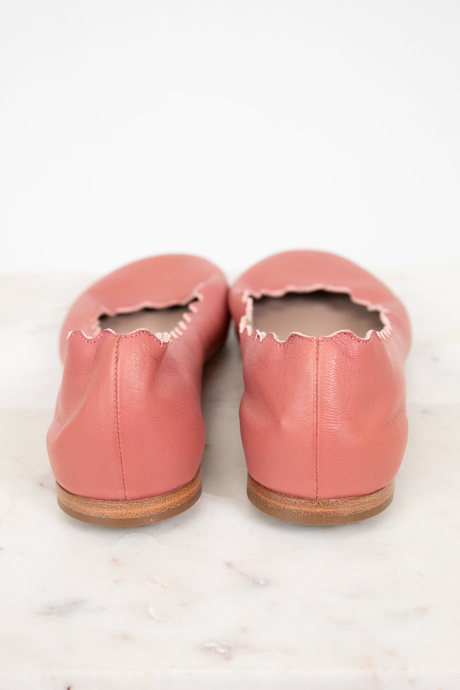 A pair of dusty pink leather ballet flats with scalloped edges, shown from the back on a white marble surface against a white background.