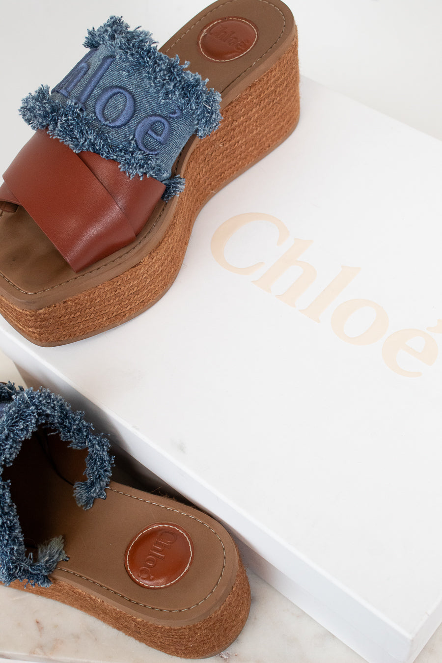 A pair of Chloé wedge sandals with brown leather straps, blue frayed trim, and jute platform espadrille soles, shown with a white Chloé shoebox.