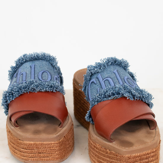 A pair of platform slide sandals featuring blue denim straps with frayed edges, embossed 'Chloé' lettering, and a brown leather strap, set on a woven jute platform sole.