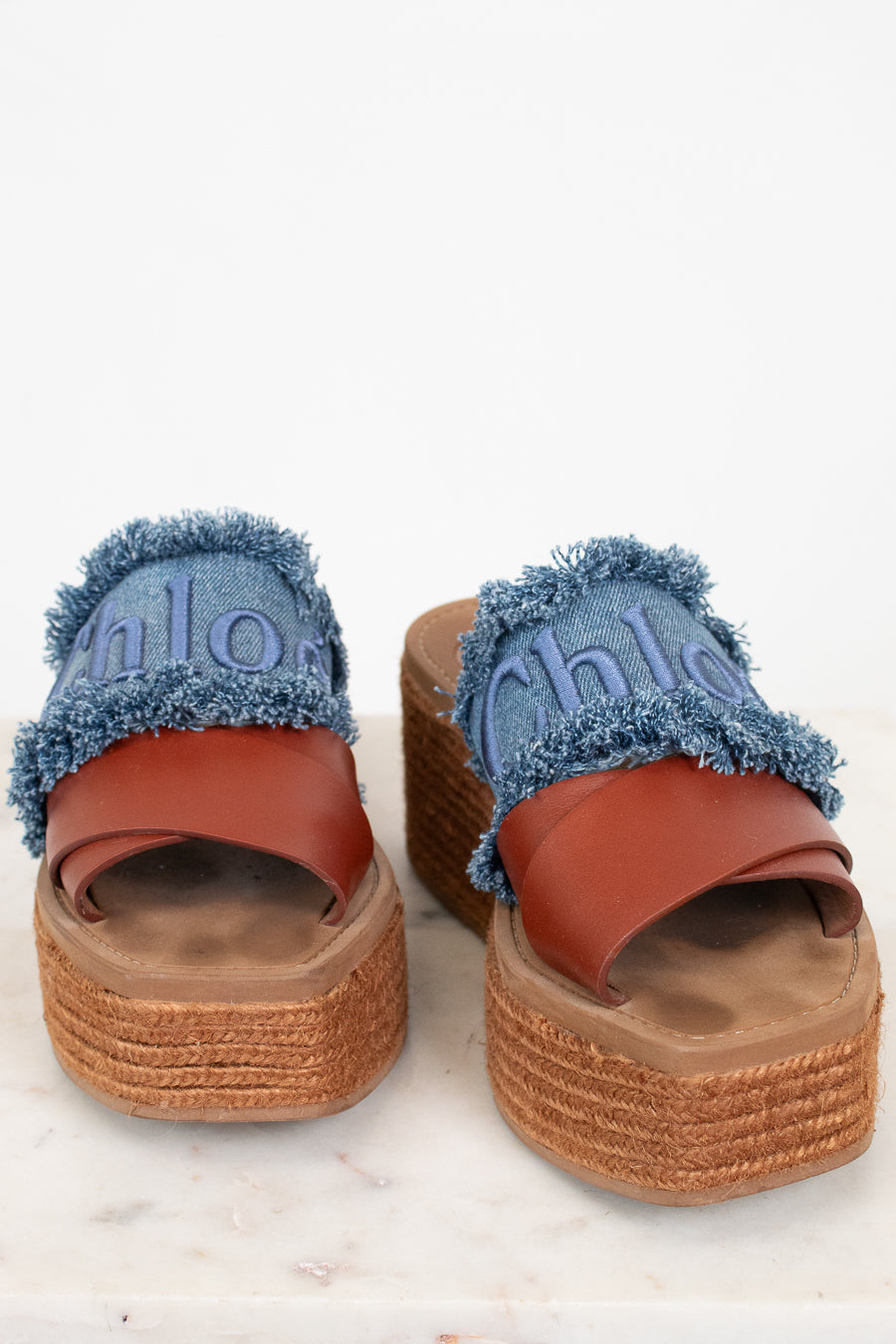 A pair of platform slide sandals featuring blue denim straps with frayed edges, embossed 'Chloé' lettering, and a brown leather strap, set on a woven jute platform sole.