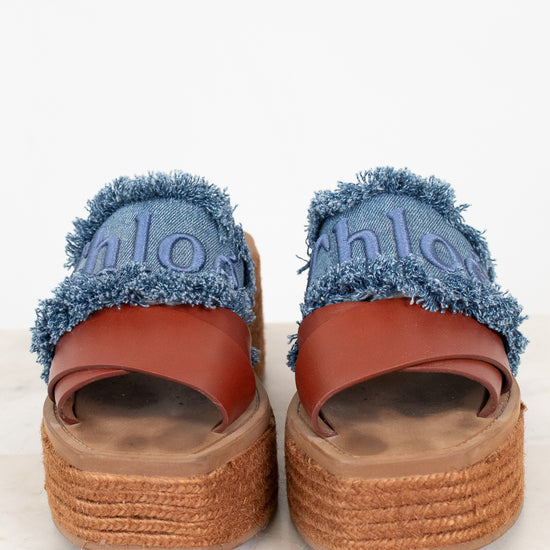 A pair of platform espadrille sandals featuring blue denim uppers with frayed edges, brown leather straps, and thick braided jute platform soles. The word 'chloé' is embroidered in blue on the denim.
