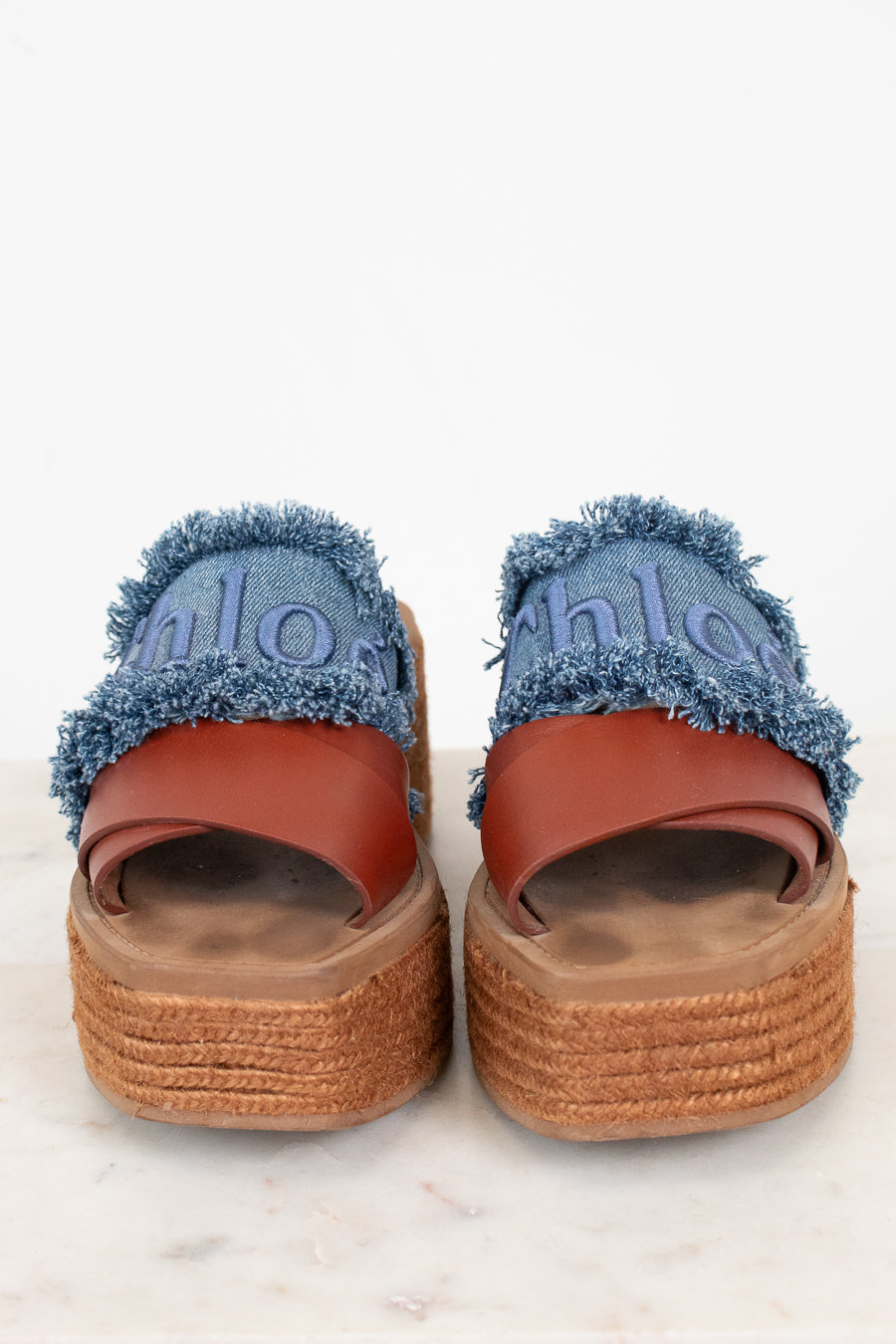 A pair of platform espadrille sandals featuring blue denim uppers with frayed edges, brown leather straps, and thick braided jute platform soles. The word 'chloé' is embroidered in blue on the denim.