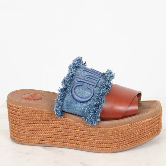 A single platform slide sandal with a brown leather strap, a blue denim fringe accent, and a brown jute-wrapped wedge sole, shown on a light marble surface against a white background.