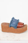 A single platform slide sandal with a brown leather strap, a blue denim fringe accent, and a brown jute-wrapped wedge sole, shown on a light marble surface against a white background.