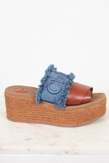 A single platform slide sandal with a brown leather strap, a blue denim fringe accent, and a brown jute-wrapped wedge sole, shown on a light marble surface against a white background.