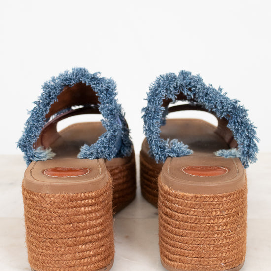 A pair of women's espadrille wedge sandals with frayed blue denim-style straps and brown woven jute platform soles, shown from the back on a light marble surface.