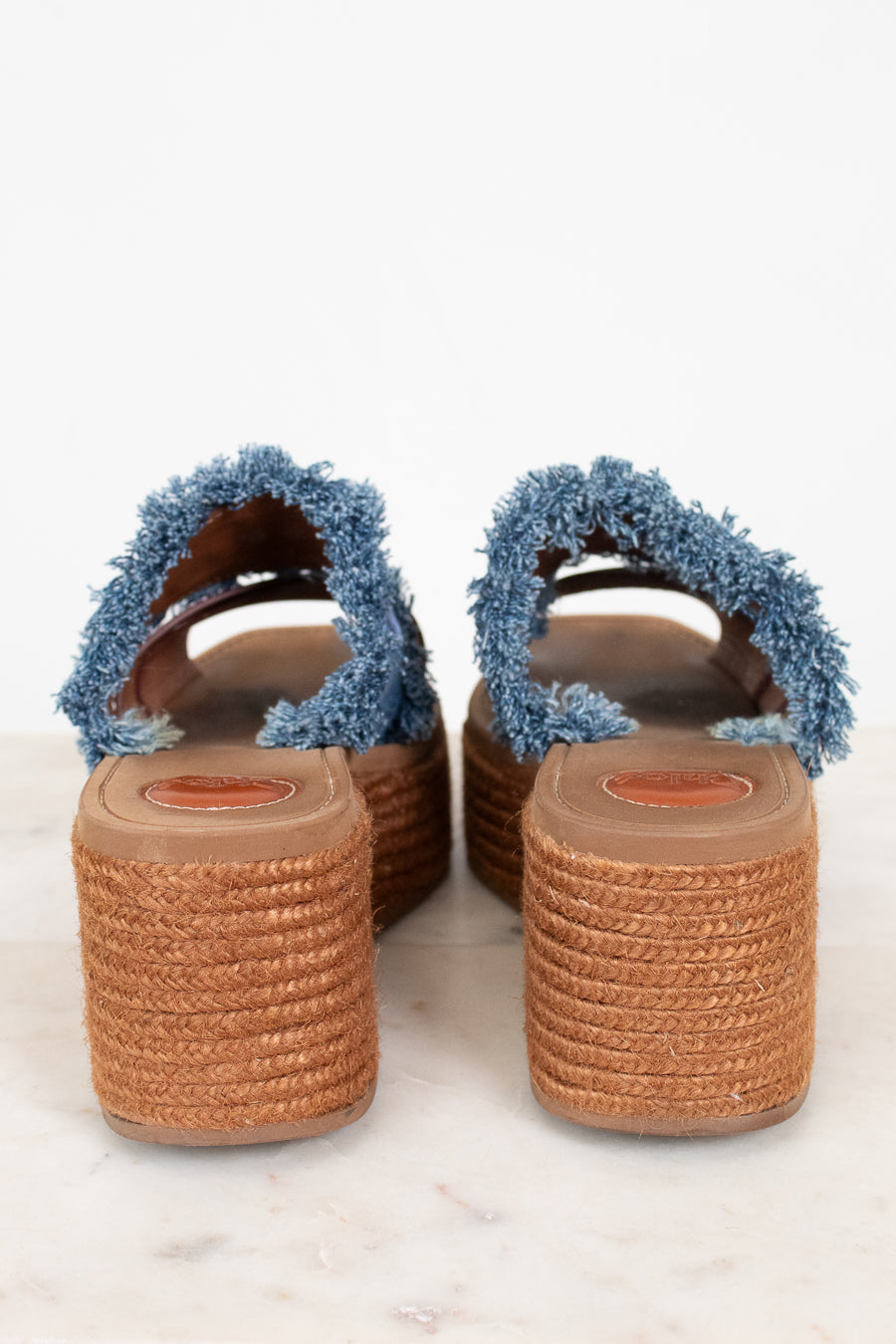 A pair of women's espadrille wedge sandals with frayed blue denim-style straps and brown woven jute platform soles, shown from the back on a light marble surface.
