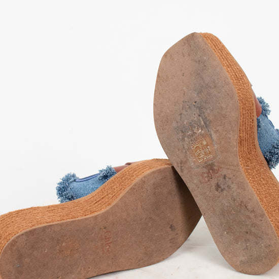 A pair of platform mule sandals shown from the bottom, displaying the brown jute-wrapped platform espadrille soles and a blue denim upper with frayed tassel details, resting on a white marble surface against a white background.