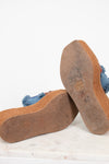 A pair of platform mule sandals shown from the bottom, displaying the brown jute-wrapped platform espadrille soles and a blue denim upper with frayed tassel details, resting on a white marble surface against a white background.