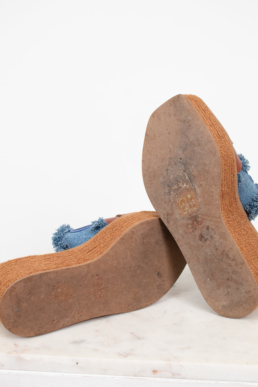 A pair of platform mule sandals shown from the bottom, displaying the brown jute-wrapped platform espadrille soles and a blue denim upper with frayed tassel details, resting on a white marble surface against a white background.