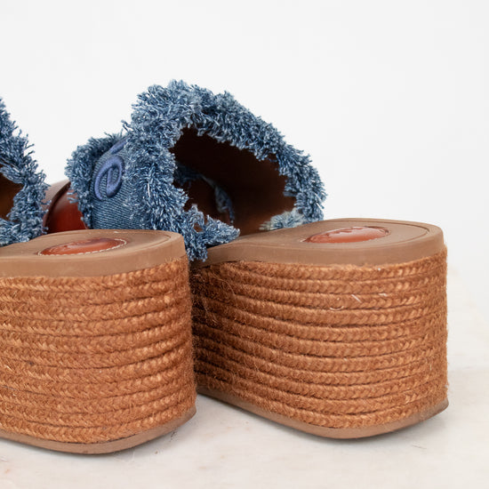 Close-up of a pair of espadrille-style wedge sandals with blue denim frayed uppers and tan braided jute platform soles on a light marble surface against a white background.