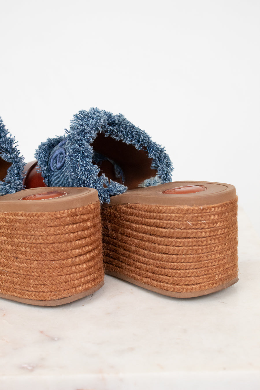 Close-up of a pair of espadrille-style wedge sandals with blue denim frayed uppers and tan braided jute platform soles on a light marble surface against a white background.