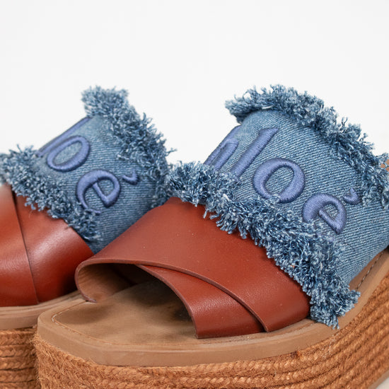 A pair of platform slide sandals featuring brown leather straps, a thick braided jute platform sole, and a frayed blue denim upper with the brand name 'Chloé' embroidered in denim.